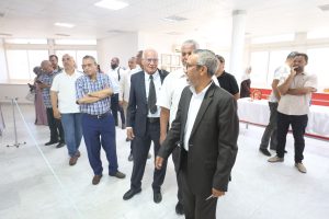 WhatsApp Image 2026-06-14 at 12.43.23 PM (2) - جامعة سبها | Sebha University A group of men in business and casual attire standing in a bright, modern room with large windows, waiting in line along a taped boundary on the floor.