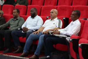 WhatsApp Image 2026-06-14 at 12.43.24 PM (2) - جامعة سبها | Sebha University Four men sit in a row of red theater seats, watching a presentation in a conference or cinema setting with attentive expressions