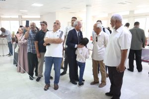 WhatsApp Image 2026-06-14 at 12.43.24 PM - جامعة سبها | Sebha University A diverse group of adults standing in a bright indoor hallway, many with smartphones raised to take photos, as they chat and wait near a white barrier line.