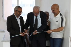 WhatsApp Image 2026-06-14 at 12.45.25 PM - جامعة سبها | Sebha University Three men in suits and a polo shirt cut a light blue ribbon at a ceremony, standing by a white wall and windowed background.