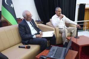 WhatsApp Image 2026-06-14 at 12.45.26 PM (2) - جامعة سبها | Sebha University Two men sit on beige sofas in a panel setting; one speaks into a handheld microphone while the other reviews documents, with a laptop nearby and a flag in the background.