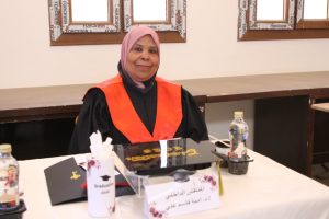 Graduate in black gown with orange stole sits at a table with a diploma and cap during a ceremony.