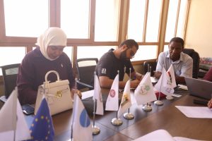 inbound1610053573953665732 - جامعة سبها | Sebha University Three people sit at a conference table signing papers, with multiple small flags along the table edge in a bright meeting room.