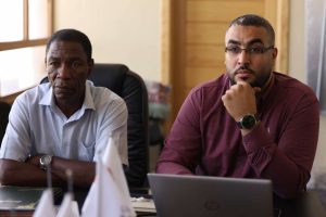 inbound2025410317778071574 - جامعة سبها | Sebha University Two men sit at a desk in an office; the man on the left wears a light shirt, the man on the right wears a maroon shirt and glasses, resting his chin on his hand with a laptop in view.
