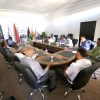 Group of professionals seated around a large conference table in a bright meeting room, with flags in the background behind the attendees.