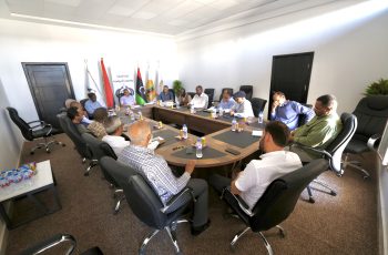 Group of professionals seated around a large conference table in a bright meeting room, with flags in the background behind the attendees.