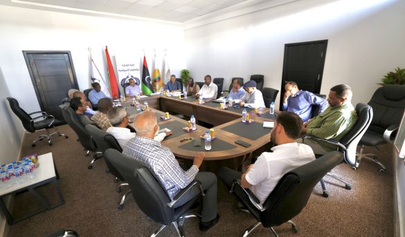 Group of professionals seated around a large conference table in a bright meeting room, with flags in the background behind the attendees.