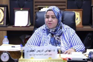Woman in blue patterned hijab and striped blouse sits at a desk during a meeting, with papers and a name plate in front.