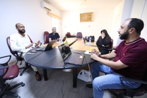 A diverse group of five people seated around a conference table, each with laptops, engaged in a discussion in a bright office.