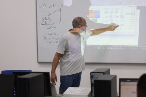 06238E51-31B3-4FCD-B2AE-4081B862E87E - جامعة سبها | Sebha University Male instructor with gray beard points at a projected slide in a classroom, whiteboard with handwritten notes in the background, computers in foreground