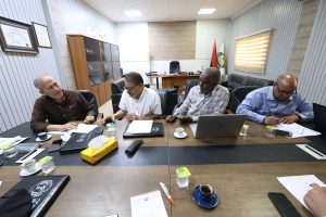 07435DD1-D83D-42BF-BEAD-9D387AA2C7F3 - جامعة سبها | Sebha University Four men sit around a long conference table in a meeting room, reviewing documents and using laptops.
