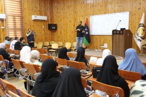 0EBD82FA-45AA-4C42-8617-FF7777A042E4 - جامعة سبها | Sebha University Speaker gesturing while presenting to an audience in a wood-paneled lecture hall with a Libyan flag beside the podium.