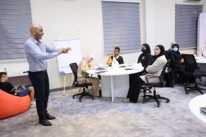 Male instructor stands by a whiteboard, teaching a diverse group of students around a round table in a classroom.