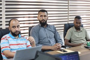 135BF045-9DB6-40D7-9971-A2FACE0EFD89 - جامعة سبها | Sebha University Three men sit at a conference table in an office; center man has a beard and wears a gray shirt, others sit nearby with laptops visible.