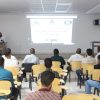 Lecturer at a desk presenting to a classroom with a projected slide and Arabic text, audience seated in rows.