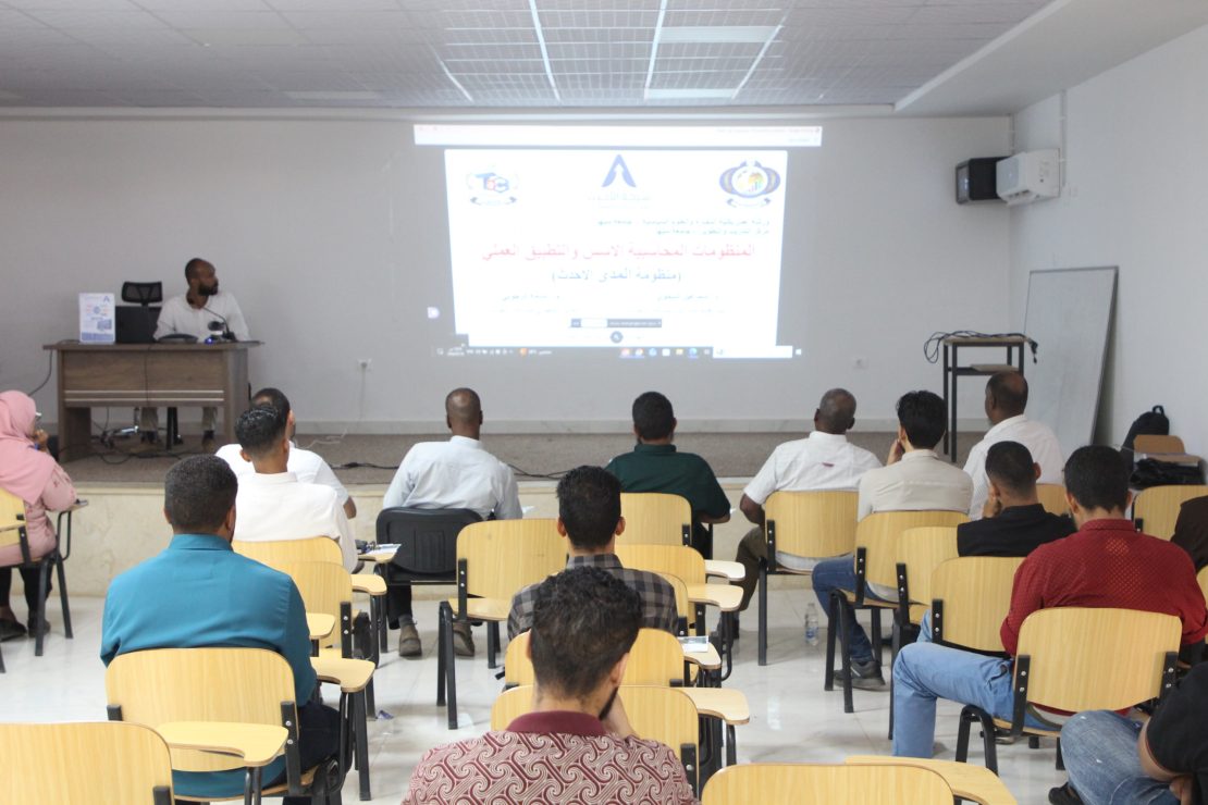 Lecturer at a desk presenting to a classroom with a projected slide and Arabic text, audience seated in rows.
