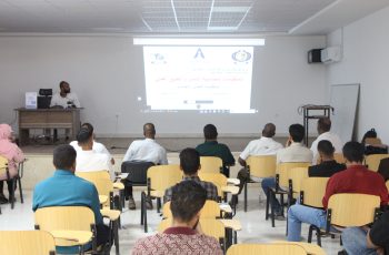 Lecturer at a desk presenting to a classroom with a projected slide and Arabic text, audience seated in rows.
