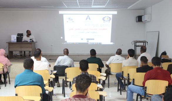 Lecturer at a desk presenting to a classroom with a projected slide and Arabic text, audience seated in rows.