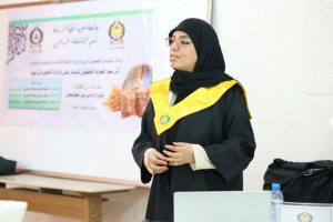 Woman in a black graduation gown and yellow stole giving a speech in a classroom or conference room.