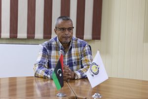 1E9EE97B-EC9B-4975-B7FF-6A4079E5D2B8 - جامعة سبها | Sebha University Middle-aged man with glasses in a plaid shirt sits at a wooden conference table, with two flags in front of him.