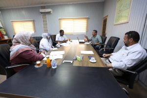 1ED579D2-2570-4207-935B-6EC20C42767A - جامعة سبها | Sebha University Group of five professionals seated around a conference table, discussing documents with laptops and beverages present.