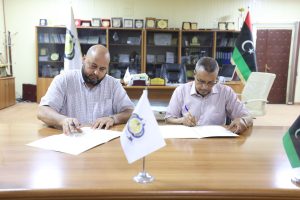 2034CEDA-C1B8-4D9A-824D-07B2973F2098 - جامعة سبها | Sebha University Two men sit at a wooden conference table signing documents in an office with awards and flags in the background.