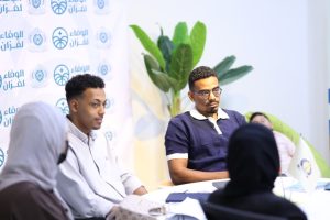 Panel discussion with two men seated at a table, backdrop logo banners and a green plant in the background.