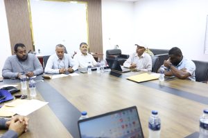 Five men in a meeting around a wooden conference table with laptops, papers, and water bottles in a bright office.