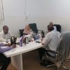 Seven people sit around a white conference table in a plain room, with tea/coffee pots, water bottles, and snacks on the table.