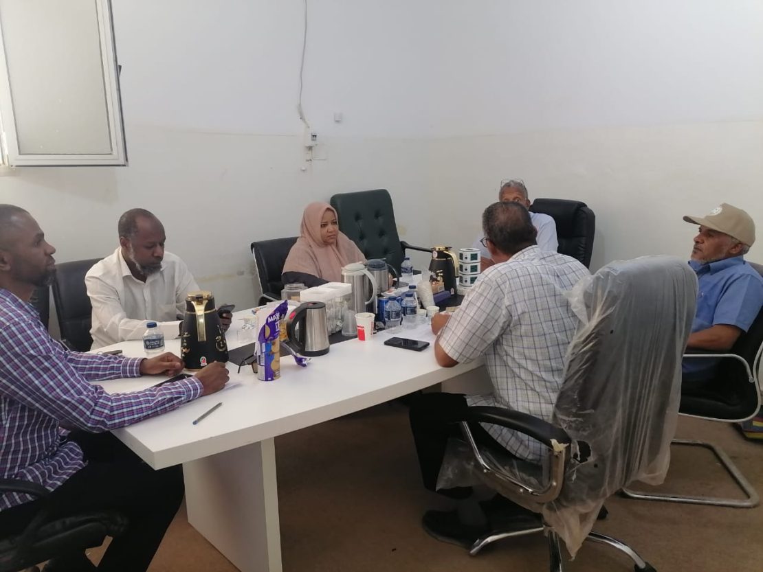 Seven people sit around a white conference table in a plain room, with tea/coffee pots, water bottles, and snacks on the table.