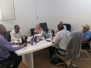Seven people sit around a white conference table in a plain room, with tea/coffee pots, water bottles, and snacks on the table.