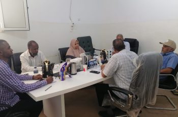Seven people sit around a white conference table in a plain room, with tea/coffee pots, water bottles, and snacks on the table.
