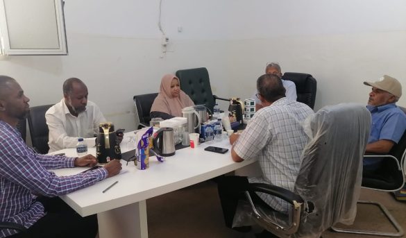 Seven people sit around a white conference table in a plain room, with tea/coffee pots, water bottles, and snacks on the table.