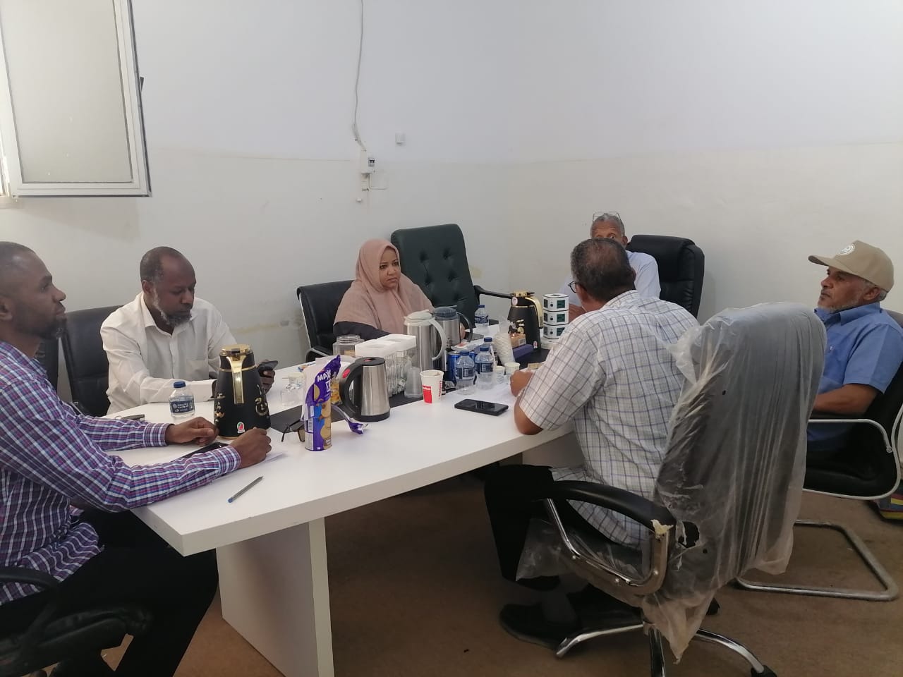 Seven people sit around a white conference table in a plain room, with tea/coffee pots, water bottles, and snacks on the table.