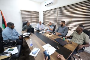 2E75545D-F3FA-4136-A7FA-066A279B9A4D - جامعة سبها | Sebha University Group of six men in a boardroom around a table with laptops and papers, one gesturing while speaking.