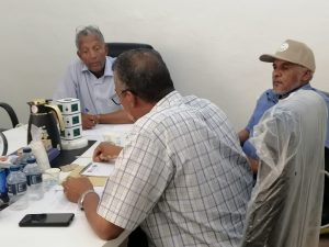 Group of three men in a meeting around a table, one writing notes while two listen attentively.