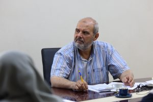 Middle-aged man with a gray beard in a blue-striped shirt sits at a conference table, writing on papers during a meeting in an office.