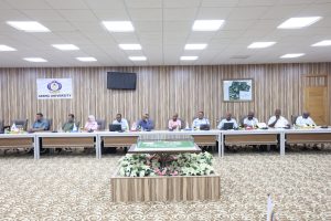 311F438F-539C-45E6-925E-7A8CE66E58D6 - جامعة سبها | Sebha University Panel of ten people seated at a long conference table in a university meeting room, with a floral centerpiece in the foreground and 'Sebha University' sign on the wall behind them