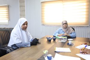 Two women in hijabs sit at a conference table in a meeting room with papers, pens, and cups scattered on the table.