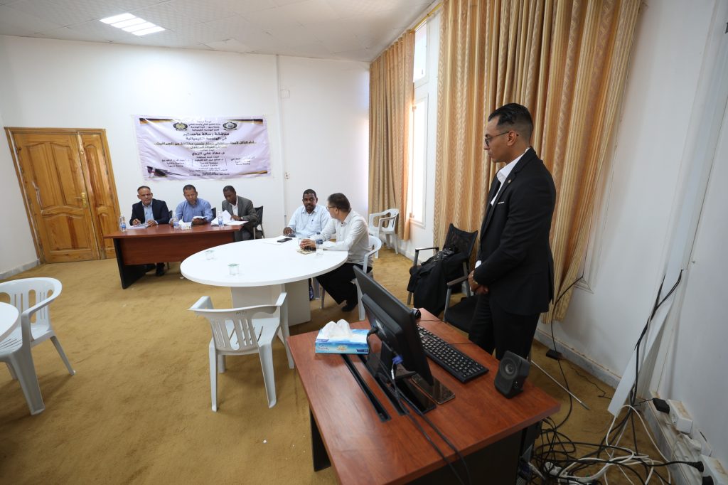 34005460-5897-4A14-AA48-5E72B3429AB0 - جامعة سبها | Sebha University Five men sit around a wooden table for a meeting, with a man in a suit standing beside a laptop in the foreground and a banner on the wall behind them