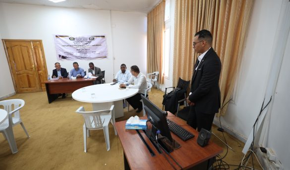 Five men sit around a wooden table for a meeting, with a man in a suit standing beside a laptop in the foreground and a banner on the wall behind them