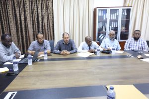 Six men sit around a large conference table in a meeting room, papers and water bottles in front of them.
