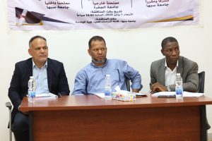 Three men in suits sit at a conference table during a meeting, with a banner in Arabic on the wall behind them.