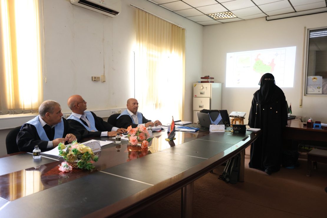 Four men in suits with light-blue sashes sit at a conference table as a woman in a black hijab presents a map on the projector screen behind them while flowers and small flags sit on the table.
