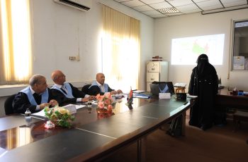 Four men in suits with light-blue sashes sit at a conference table as a woman in a black hijab presents a map on the projector screen behind them while flowers and small flags sit on the table.