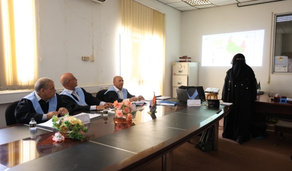 37F5E589-C96D-46FD-9107-983C2F2FCC38 - جامعة سبها | Sebha University Four men in suits with light-blue sashes sit at a conference table as a woman in a black hijab presents a map on the projector screen behind them while flowers and small flags sit on the table.