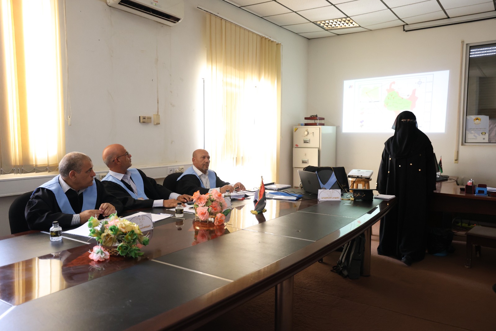 Four men in suits with light-blue sashes sit at a conference table as a woman in a black hijab presents a map on the projector screen behind them while flowers and small flags sit on the table.