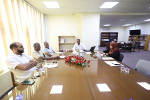 3AB55B8B-E229-453B-A8E5-B4E70F1E0C54 - جامعة سبها | Sebha University Five people sit around a long conference table in a bright office, discussing with papers, laptops, and cups; a red flower centerpiece dominates the table.
