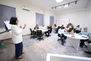 Woman presenter in a white blazer speaks to a diverse group at round tables in a bright training room with orange flags on the tables.
