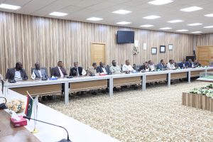 49858935-BA91-4B77-BDF7-EE95FF0EAE72 - جامعة سبها | Sebha University Group of professionals in suits seated along a long conference table in a wood-paneled room, with microphones, documents, and small flags on the table.